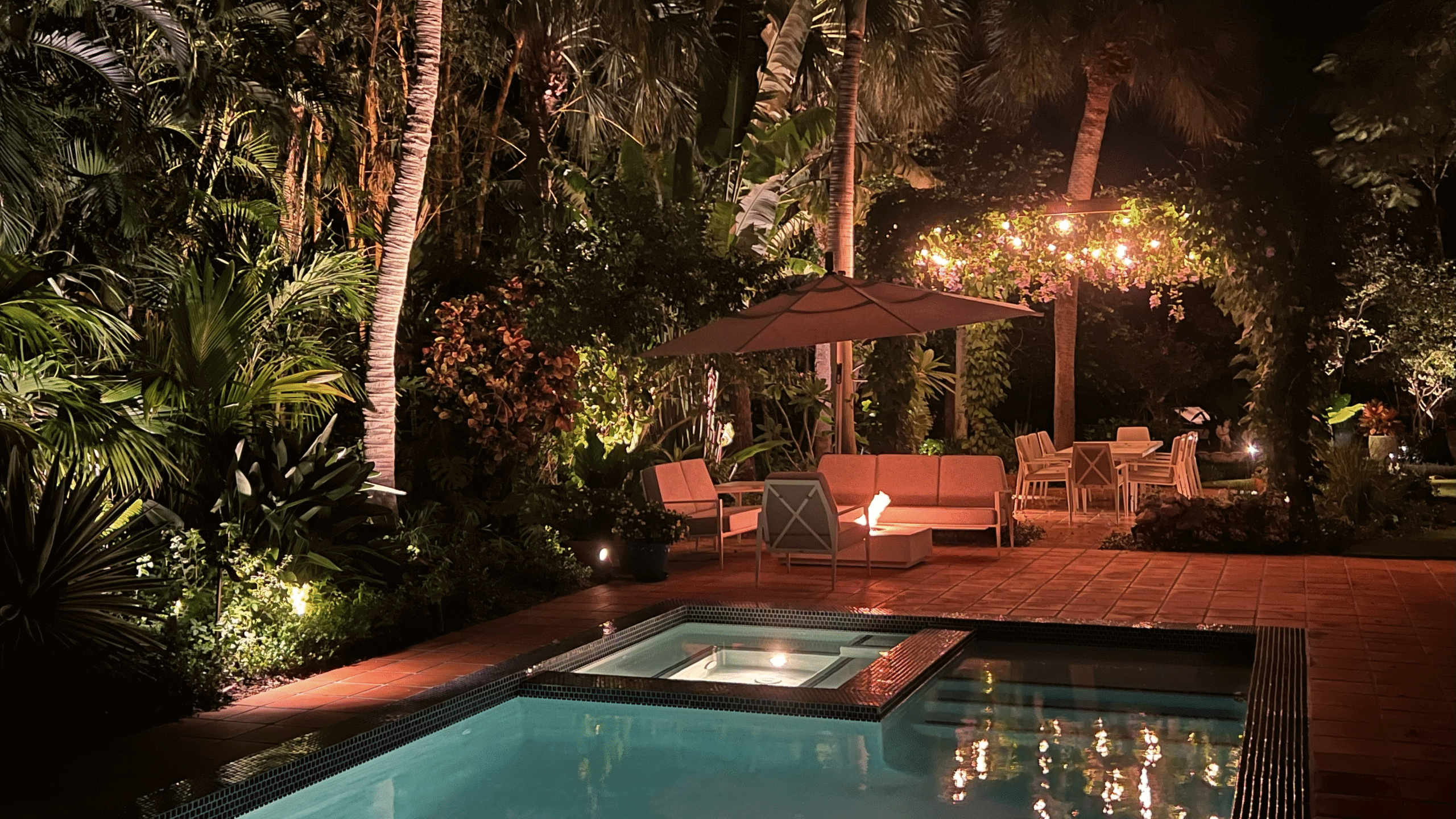 Nighttime pool and patio with lush greenery.